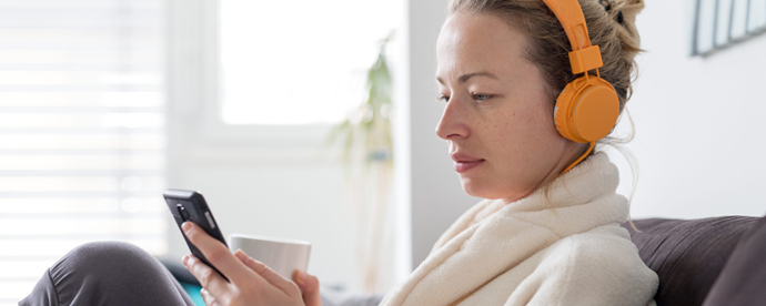 Kvinna med hörlurar tittar på sin telefon och har en kaffekopp i handen